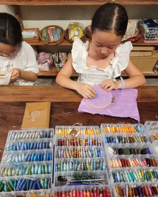 A child embroiders purple fabric at a table with boxes of coloured thread