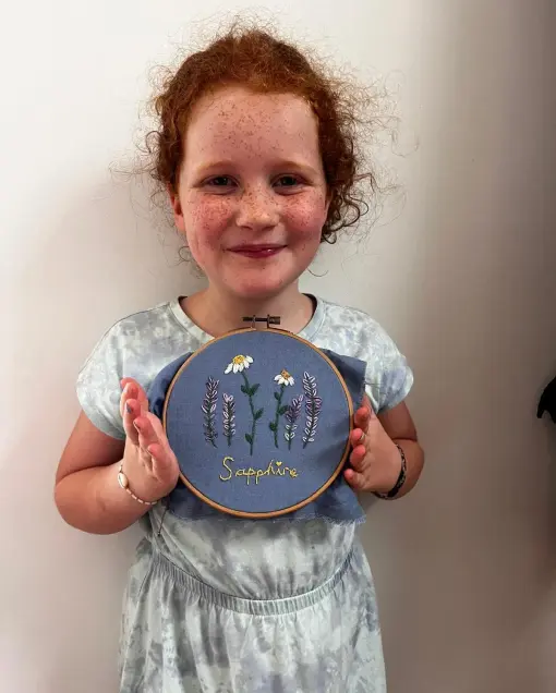 A child holds a finished embroidery hoop with daisies and lavender on blue fabric