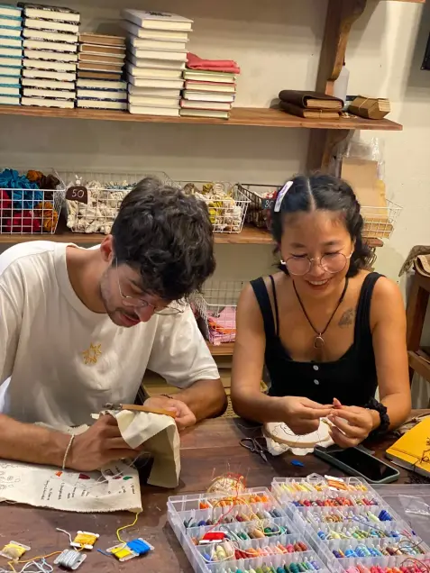 Two guests embroider at a table with a box of coloured thread
