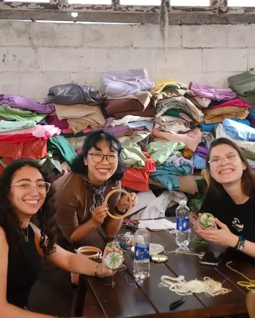 Three people hold embroidery hoops in front of a pile of coloured fabric